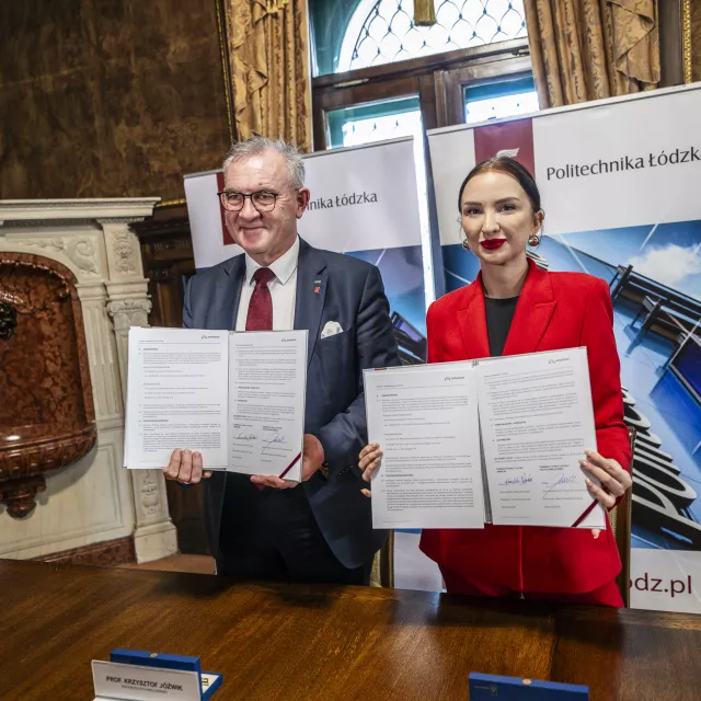 Kobieta w czerwonym garniturze i mężczyzna w garniturze pokazują, stojąc za stołem, podpisany dokument