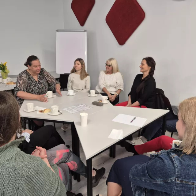 Grupa osób siedzi przy stole prowadząc rozmowę. Stół ma nieregularny kształt i stoja na nim filiżanki