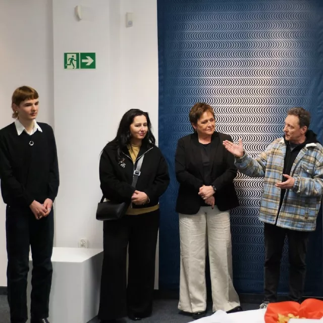 Stoi grypa osób, jedna z nich gestykuluje rekoma, wygląda jakby przedstawiała jedną z osób.